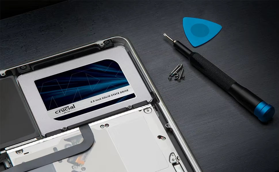 A silver Crucial 2.5-inch solid state drive installed in a laptop, shown alongside a blue plastic opening tool, a black and blue precision screwdriver, and small screws on a dark textured surface.
