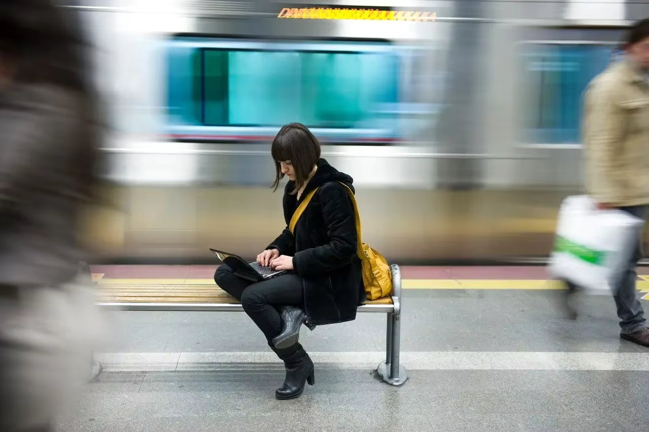 A woman sits on a subway bench using her phone, with a blurred train and other commuters in a city station. She is wearing a black coat, black pants, and black boots, and has a bright yellow shoulder bag.