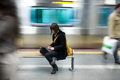 A woman sits on a subway bench using her phone, with a blurred train and other commuters in a city station. She is wearing a black coat, black pants, and black boots, and has a bright yellow shoulder bag.