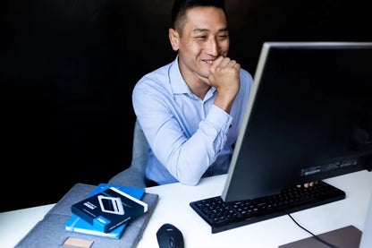 A man in a light blue dress shirt sits at a desk in front of a computer monitor, with a keyboard, mouse, and stacked notebooks on the table.